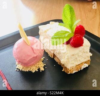 A slice of white chocolate cheesecake garnished with raspberries and mint leaves. A ball of raspberry sorbet with a white chocolate shard. Stock Photo
