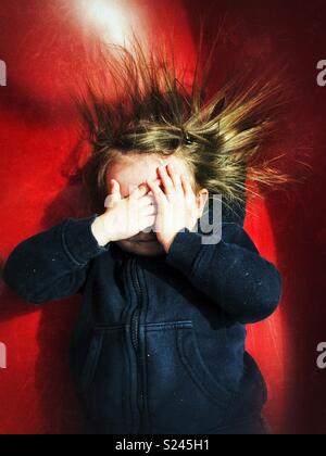 Toddler girl covering face with hands and hair sticking up due to static electricity from a red slide Stock Photo