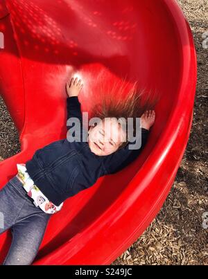 Action image of toddler girl sliding down red slide with arms up and hair sticking up Stock Photo