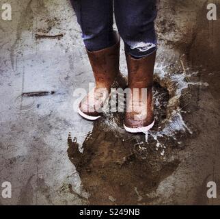 Child’s feet wearing brown rain boots jumping and splashing in a muddy puddle Stock Photo