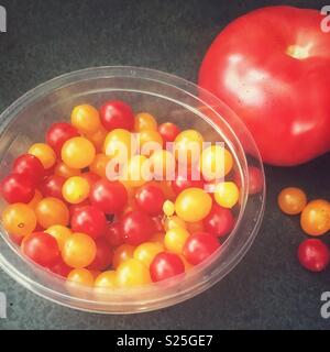 Tomberry tomatoes in a clear plastic bowl beside a regular sized tomato to illustrate their small size Stock Photo