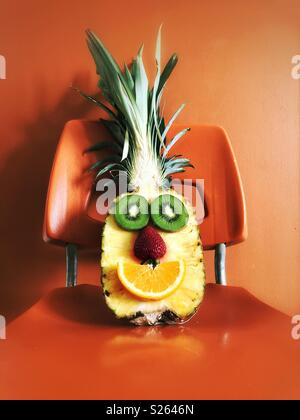 Half pineapple with kiwi slices, strawberry and orange slice made to look like a happy face sitting on an orange chair Stock Photo