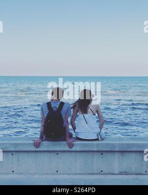 Couple sat together on a wall looking out at the sea. Stock Photo