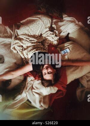 Teen girl lying on blankets and pillows with arms open and eyes closed as if she’s taking a break from her smartphone Stock Photo