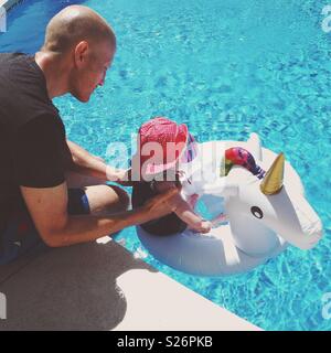 Father putting baby in inflatable unicorn in the pool Stock Photo