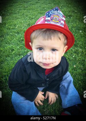 Little boy sitting in grass wearing fireman helmet, vignette filter Stock Photo