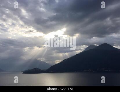 Sun rays breaking through clouds at sunrise with lake and mountains Stock Photo