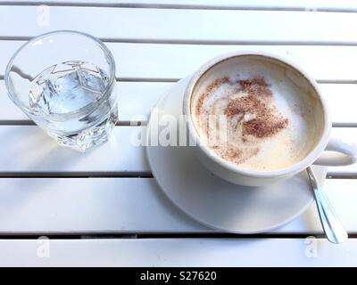 A cup of cappuccino coffee and a glass of water on a white wooden, slatted table Stock Photo