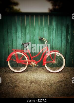 Vintage red Flyer bicycle with white tires leaning against a green wooden fence on a gravel path.