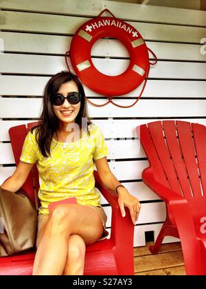 Beaming smiling pretty young brunette woman with sunglasses and short shorts sitting legs crossed in red Adirondack deck chair Stock Photo