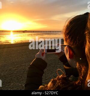 Young girl taking photo of sunset over the sea a on mobile phone. Stock Photo