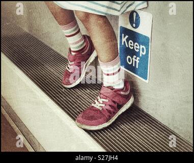 A girl rests her feet on a ledge that is also a radiator. The sign says “Keep Off!” She’s ignoring the request. Children never listen?! Photo Credit - © COLIN HOSKINS. Stock Photo