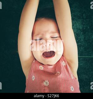 Toddler girl having a temper tantrum with arms up and mouth open screaming on a tile floor Stock Photo