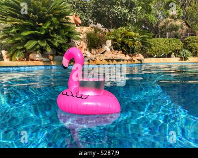 Pink flamingo drinks holder floating in a swimming pool with a glass containing an ice cold drink. Stock Photo