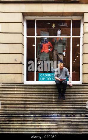 A hipster male perched on a high shop window ledge whilst looking at his mobile phone Stock Photo