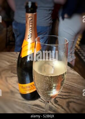 A Bottle of prosecco with a half full glass next to it in a pub with a man and a woman blurred in the background Stock Photo
