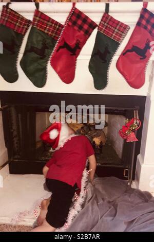 Little boy dressed in a red shirt and Santa hat looking up the chimney, waiting for Santa Claus on Christmas Eve night Stock Photo