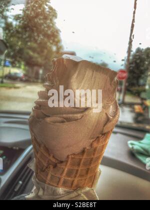 Chocolate soft serve ice cream in a waffle cone.  It is being enjoyed on a rainy day in a car. Rain drops are on the windshield, and a stop sign can be seen in the distance. Stock Photo