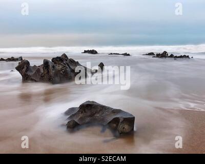 Incoming morning tide washing around rocks on a Welsh surf beach during rough weather, November. Stock Photo