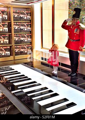 Cute little blonde girl in a red dress saluting along with FAO Schwartz soldier at the Rockefeller Center store, New York City, United States Stock Photo