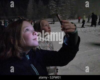 Two girls watch a nighttime light show on a ski hill. One records with her smartphone and is illuminated by the display. Stock Photo