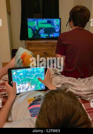Two brothers play Fortnite on their own device. The older brother is playing on the television in the background and the younger brother is playing on his tablet in the foreground. Stock Photo