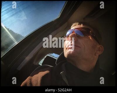 Man in the driving seat of a car with Sun on the window and a clear sky, Spain. Stock Photo