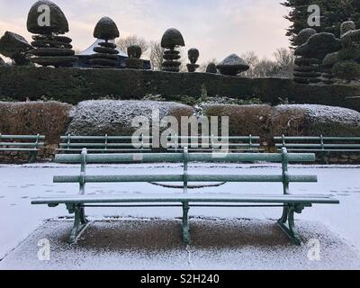 Empty park bench covered in light snow with topiary in the background, Alexandra park, Penarth, South Wales. Stock Photo