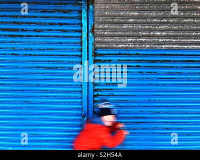 Boy on a scooter passing by a blue roller shutter. Stock Photo