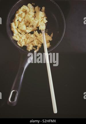 Scrambled eggs in black frying pan with wooden spoon on black stovetop Stock Photo