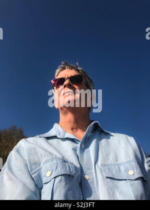 Middle aged grey haired man wearing sunglasses in a chambray shirt against a deep blue sky. Stock Photo