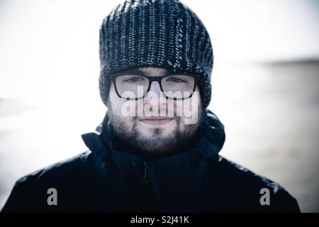 A young man with beard and glasses wearing a warm woolly hat and warm coat looking directly into the camera like an explorer or outdoor adventurer Stock Photo