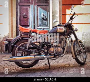 A vintage Royal Enfield motorbike parked on a cobblestone pavement in front of the entrance of a building. The entrance comprises a wooden door Stock Photo