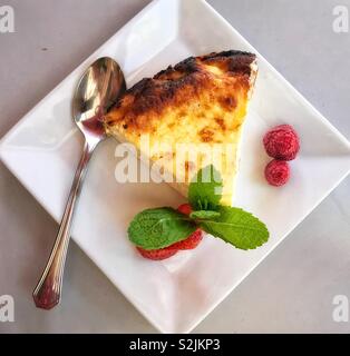 Cheesecake served in a white rectangular china dish, with  raspberries, strawberries and mint for decorative purposes and a silver plated teaspoon for the eating thereof. Stock Photo