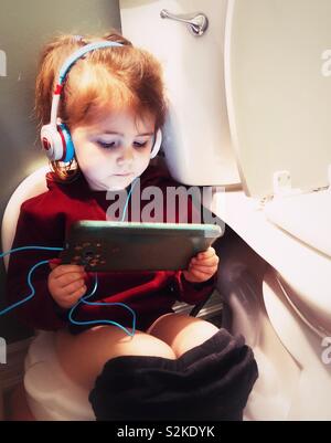 Toddler girl string on potty with tablet and headphones in a bathroom Stock Photo