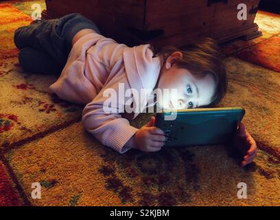 Young girl laying on floor holding a tablet that is illuminating her face Stock Photo