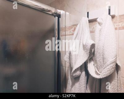 A couple of white bathrobes hanging on the wall near the glass door of a shower stall Stock Photo