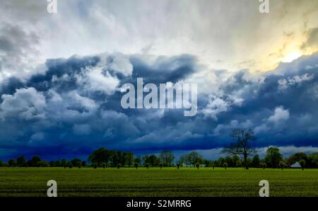 Sun breaking through behind and above the dramatic and dark clouds over a green Grassy Countryside landscape Stock Photo