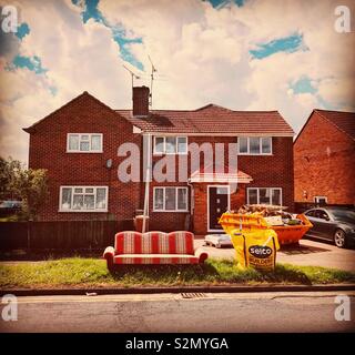 A house undergoing refurbishment with a skip on the drive and an old sofa and bag of building materials on the grass verge. Stock Photo