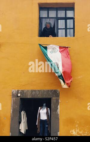 An elderly woman looks down onto the Mexican flag from her home above a clothing boutique in San Miguel de Allende, Mexico. Stock Photo