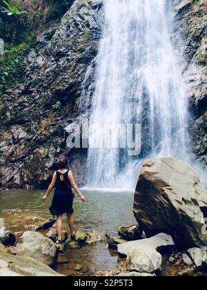 Young woman dressed in black dress enjoying the view of beautiful tropical waterfall, Thailand Stock Photo