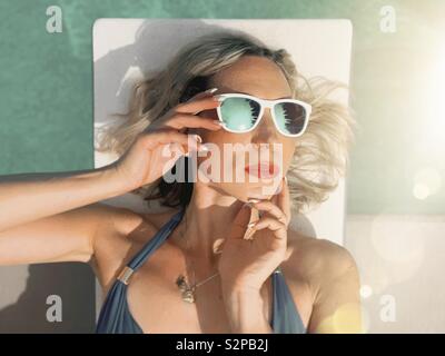 Woman wearing sunglasses lying down on white diving board with water in the background. Stock Photo