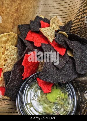 Variety of red, blue, and white corn chips set alongside a small tub of fresh green guacamole. Stock Photo