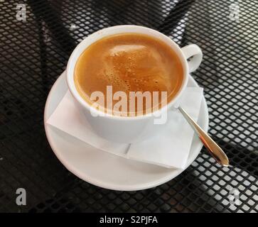 Cup of hot coffee in a while cup and saucer on a black metal mesh table top in an outdoor cafe Stock Photo