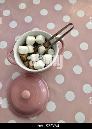 Brown and white sugar cubes and sugar tongs in a pink bowl on a pink and white spotted background Stock Photo