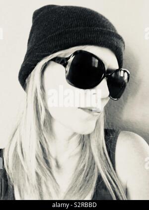 A black and white monochrome picture of a beautiful blonde hair girl wearing a beanie hat with dark sunglasses Stock Photo