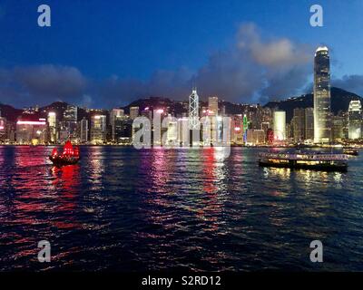 HONG KONG -29 JUN 2019- Night view of colored neon lights on tall buildings on the Hong Kong skyline with boats crossing Victoria Harbour. Stock Photo
