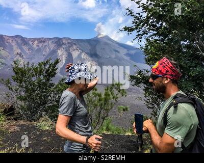 Two hikers on Mount Etna, near Catania, Sicily. The crater is active. Old lava flows are in lighter shade of grey, newer flows are darker. Blue sky day. Stock Photo