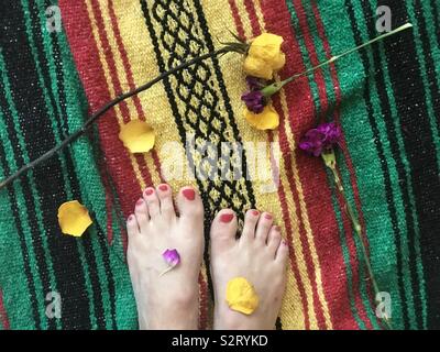 Beautiful woman’s feet with rose petals and flowers on a colorful, festive blanket. Feet and flowers Stock Photo