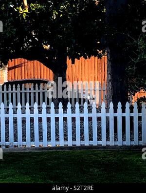 A quiet evening sunset reflects on a fence behind a white picket fence. This is suburban living at its best. Stock Photo